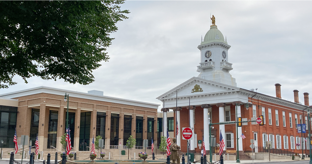 A historic brick building with a clock tower stands beside a more modern structure. American flags line the street in front, with trees partially visible on the left.