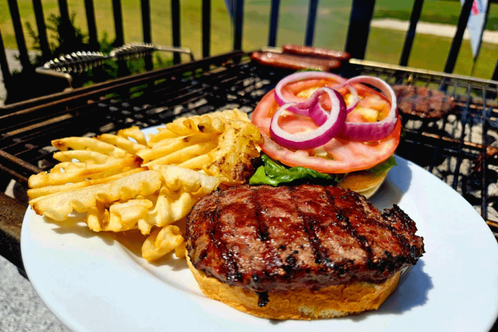 An open-face burger with onions, tomatoes, and lettuce and waffle fries on a plate next to a grill outside.