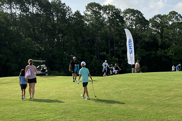 Children with golf equipment walk on the course with adults nearby
