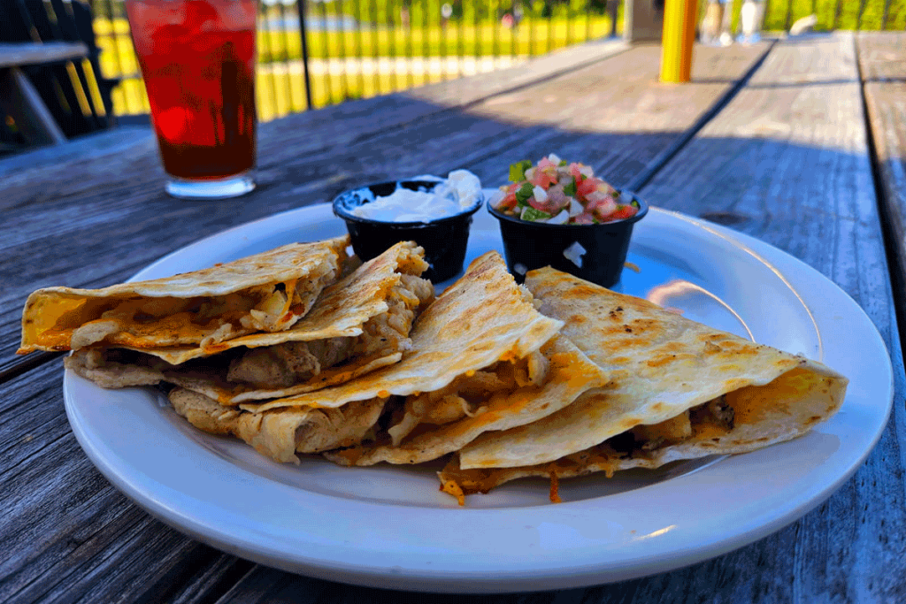 A chicken quesadilla on a plate with sour cream and salsa, on a wooden table outside in the shade,